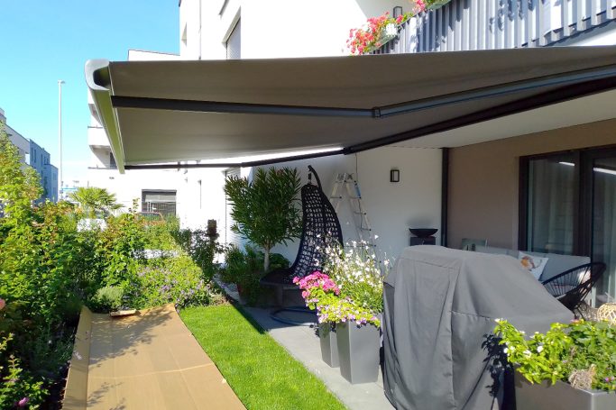 Terrasse mit Sonnenschutz, Pflanzen und Grill, blauer Himmel im Hintergrund.