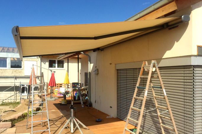Markise über einer Terrasse mit Leitern und bunten Sonnenschirmen im Hintergrund.